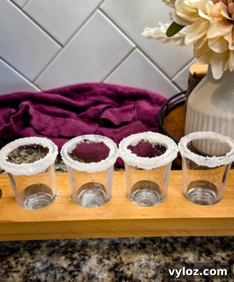 Four empty shot glasses with sugared rims lined up on a wooden tray, set on a granite countertop with a flower vase in the background.