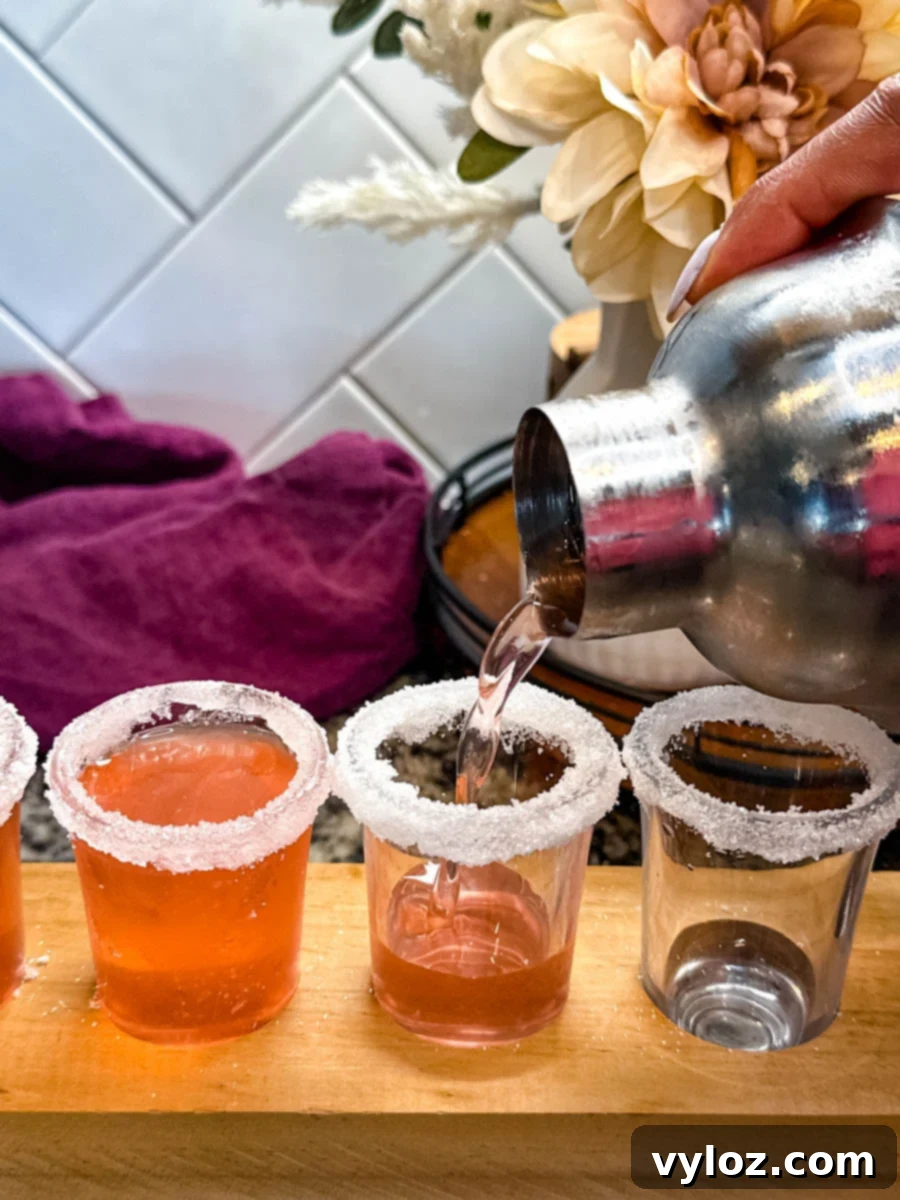 Cocktail being poured from a shaker into a sugar-rimmed shot glass, surrounded by filled glasses on a wooden tray.