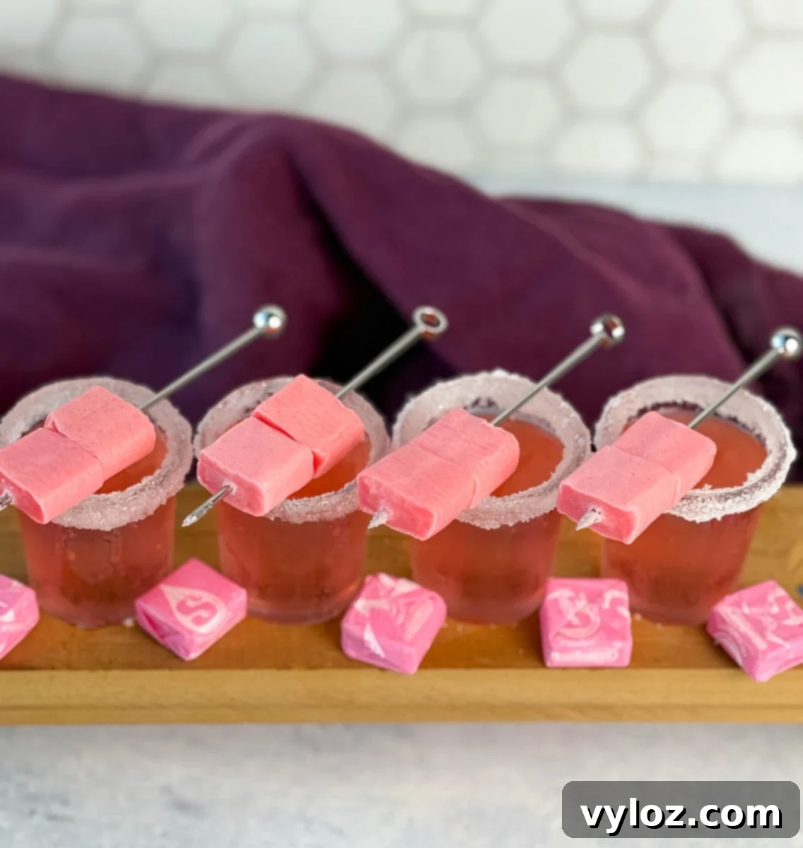 Overhead view of four pink Starburst shots with sugared rims and skewered pink Starbursts, surrounded by wrapped candy.