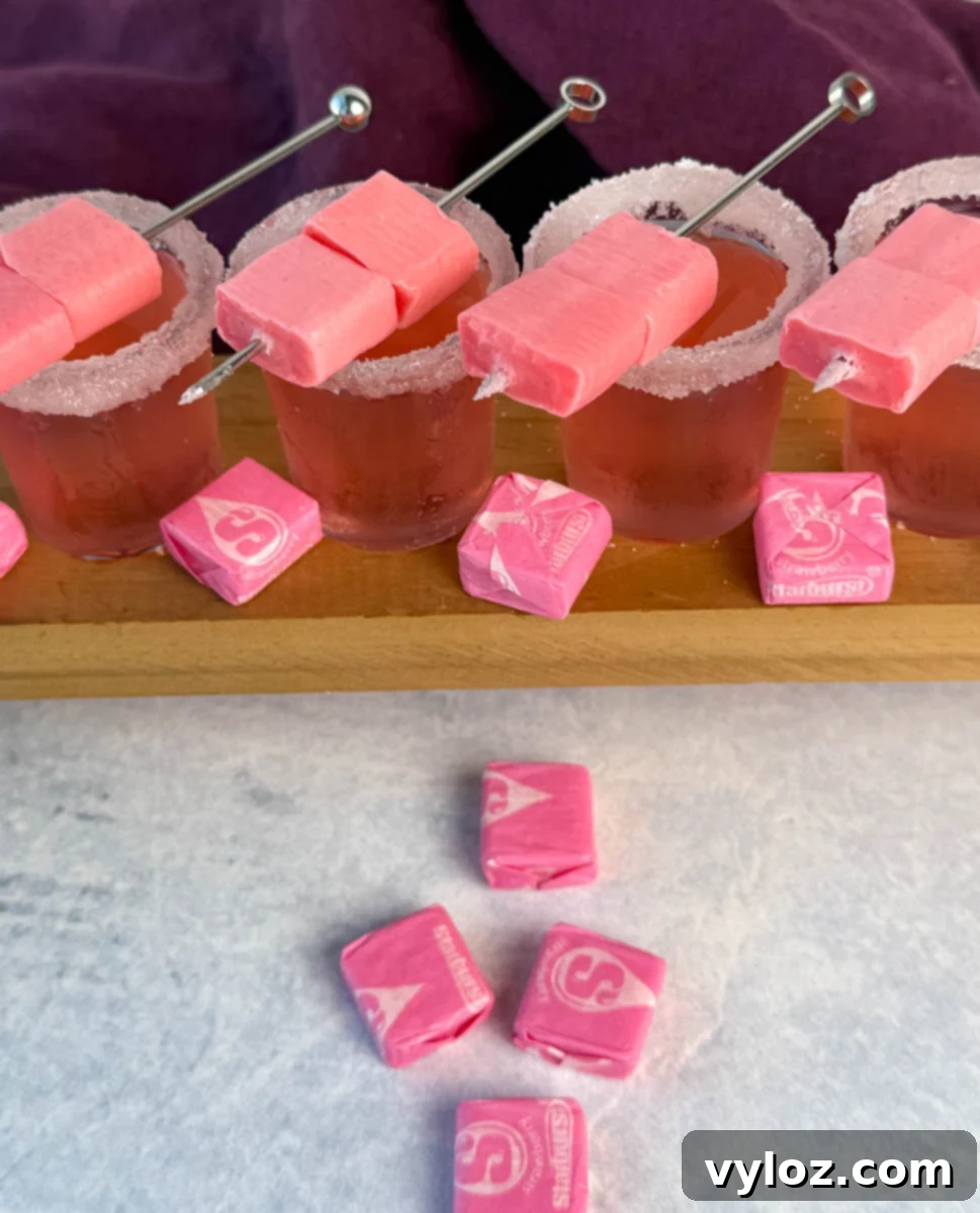 Overhead view of four pink Starburst shots with sugared rims and skewered pink Starbursts, surrounded by wrapped candy.