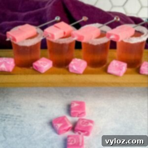 Four pink Starburst-inspired cocktail shots with sugar-rimmed glasses, each garnished with skewered pink Starburst candies. More pink Starbursts are scattered on the table in front of the drinks, with a hexagon-tiled wall and deep purple cloth in the background.