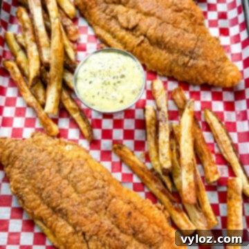 Golden fried catfish served with hand-cut fries and a side of creamy tartar sauce, all arranged on red and white checkered paper in a black tray.