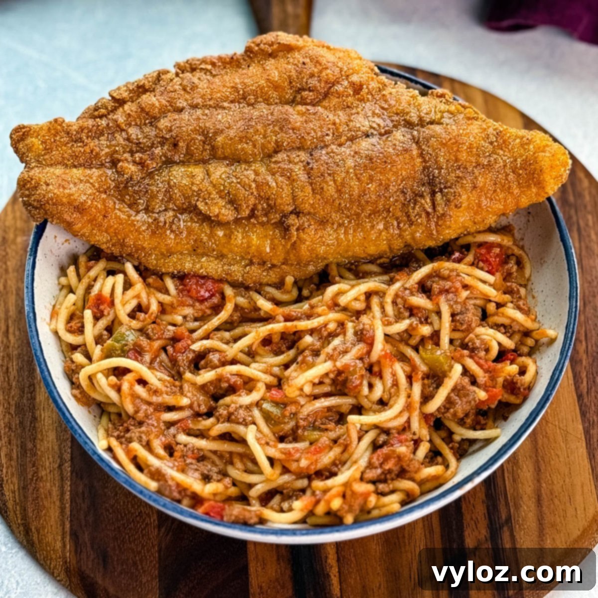 Crispy fried catfish served alongside a generous portion of beefy spaghetti, garnished with shredded cheese, on a white plate placed on a rustic wooden board.