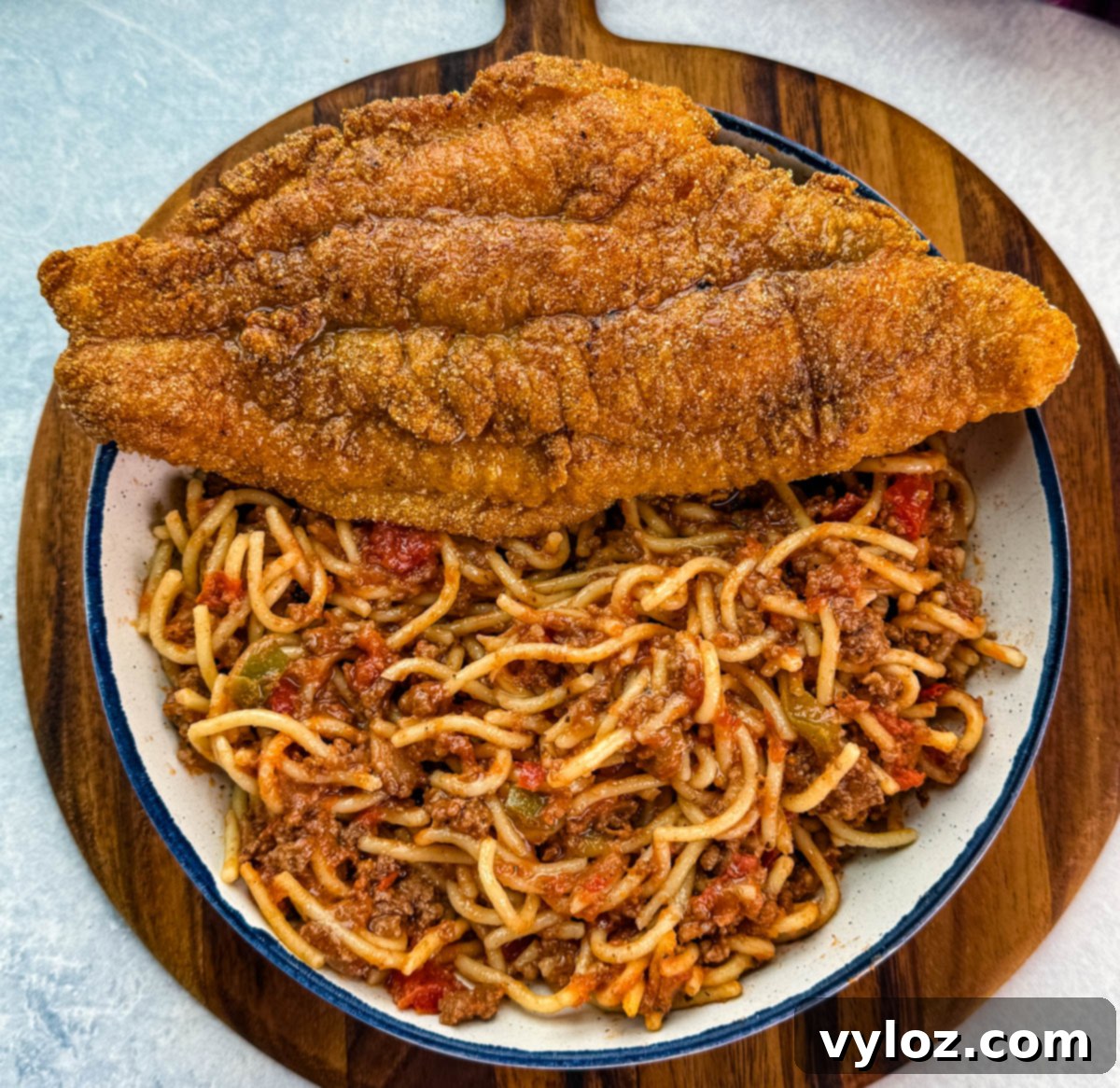 Overhead view of a beautifully presented serving of crispy fried catfish placed over a generous bowl of rich spaghetti meat sauce, all displayed on a rustic wooden cutting board.