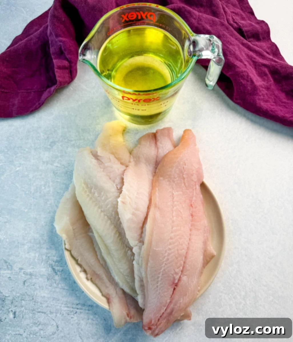 A small plate with raw catfish fillets stacked neatly, next to a glass measuring cup filled with cooking oil. A deep purple cloth napkin rests in the background on a light blue surface, ready for recipe preparation.
