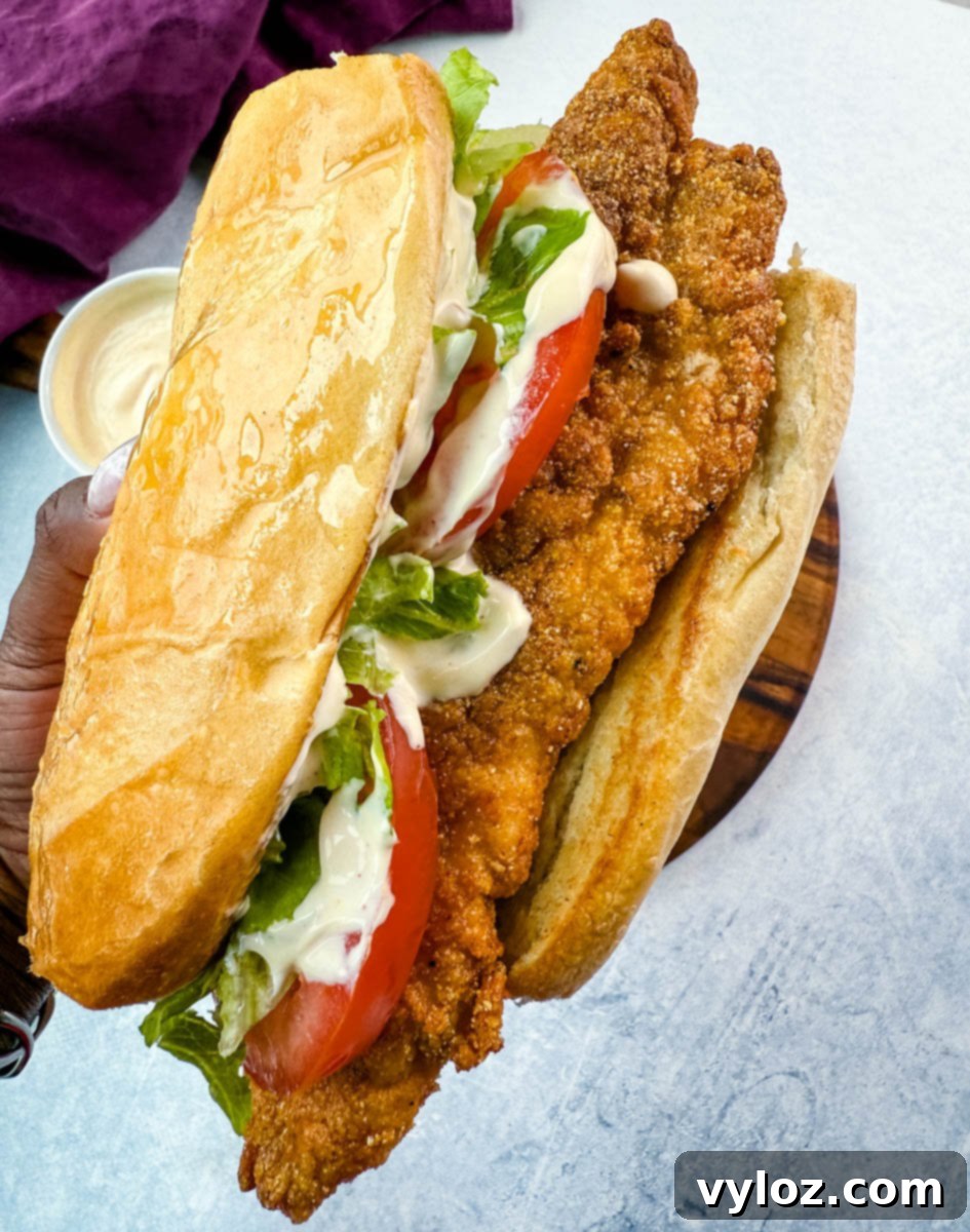 Overhead view of po' boy sandwich ingredients arranged on a round wooden cutting board. The setup includes shredded lettuce, three slices of ripe tomato, a small bowl of creamy sauce, and a large loaf of French bread. A purple cloth napkin is placed above the board on a light background.