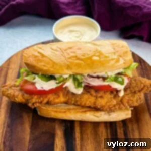 Close-up of a finished catfish po’ boy sitting on a wooden cutting board. The sandwich is stuffed with crispy golden fish, lettuce, tomato, and drizzled with creamy sauce. A bowl of extra sauce is blurred in the background.