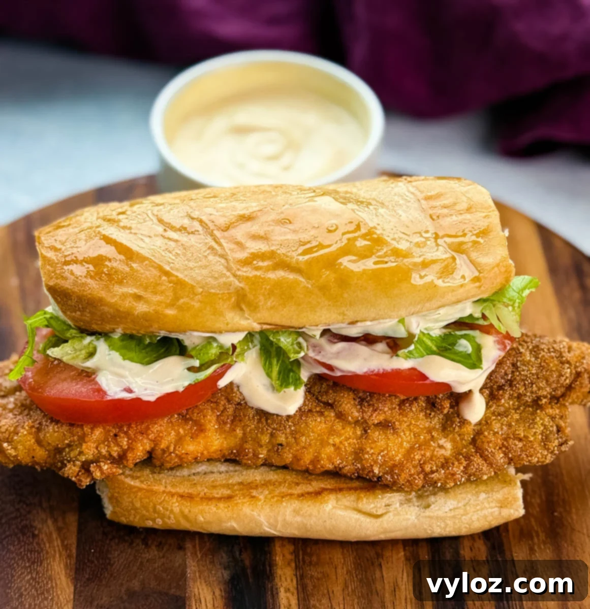 Close-up of a finished catfish po’ boy sitting on a wooden cutting board. The sandwich is stuffed with crispy golden fish, lettuce, tomato, and drizzled with creamy sauce. A bowl of extra sauce is blurred in the background.