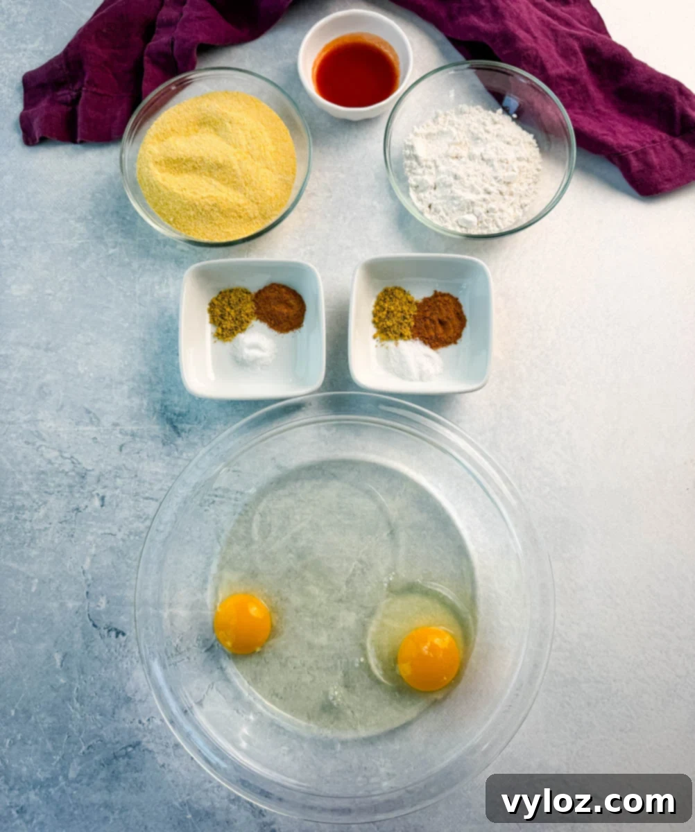 Flat lay of fish fry ingredients: cornmeal, flour, hot sauce, spices, and two cracked eggs in a clear bowl, all arranged on a light blue surface with a purple napkin.