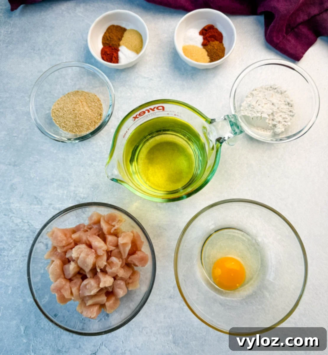 Crispy Chicken Po Boy 4 Overhead view of individual ingredients for fried chicken: chopped raw chicken, an egg in a bowl, a pile of flour, a pile of breadcrumbs, various seasonings in small dishes, and a bottle of oil. All neatly arranged for cooking preparation.
