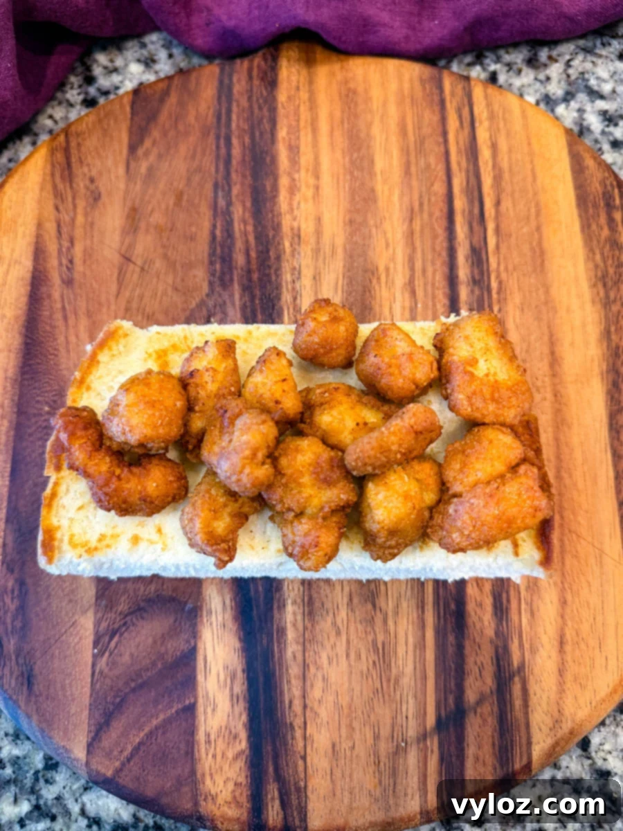 Crispy Chicken Po Boy 9 Top-down image showing the bottom half of a sandwich roll loaded with golden, crispy chicken pieces on a wooden board with a granite countertop background. Ready for toppings.