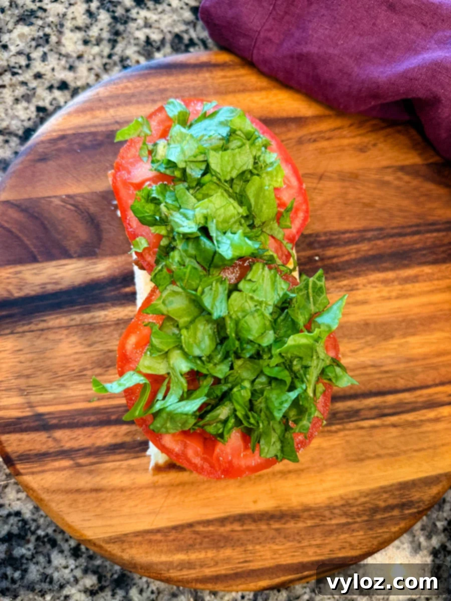 Crispy Chicken Po Boy 10 Overhead shot of a sandwich roll topped with fresh tomato slices and shredded romaine lettuce, ready for the top bun and serving. On a wooden board with a purple napkin nearby.
