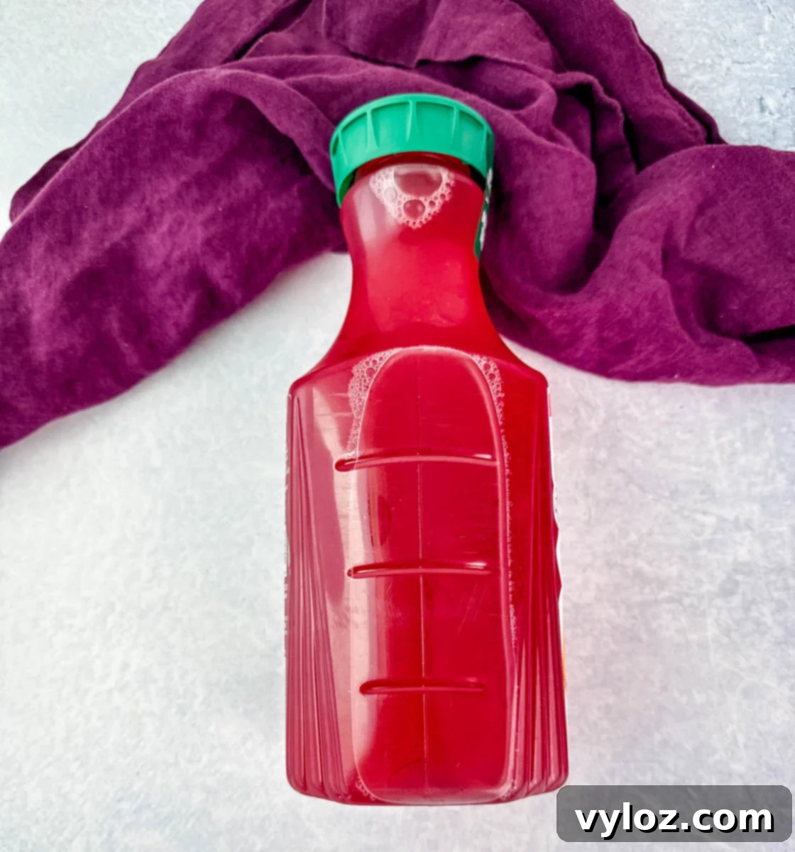 A full, unopened bottle of bright red fruit punch with a green cap, placed against a light surface and a deep purple cloth backdrop.