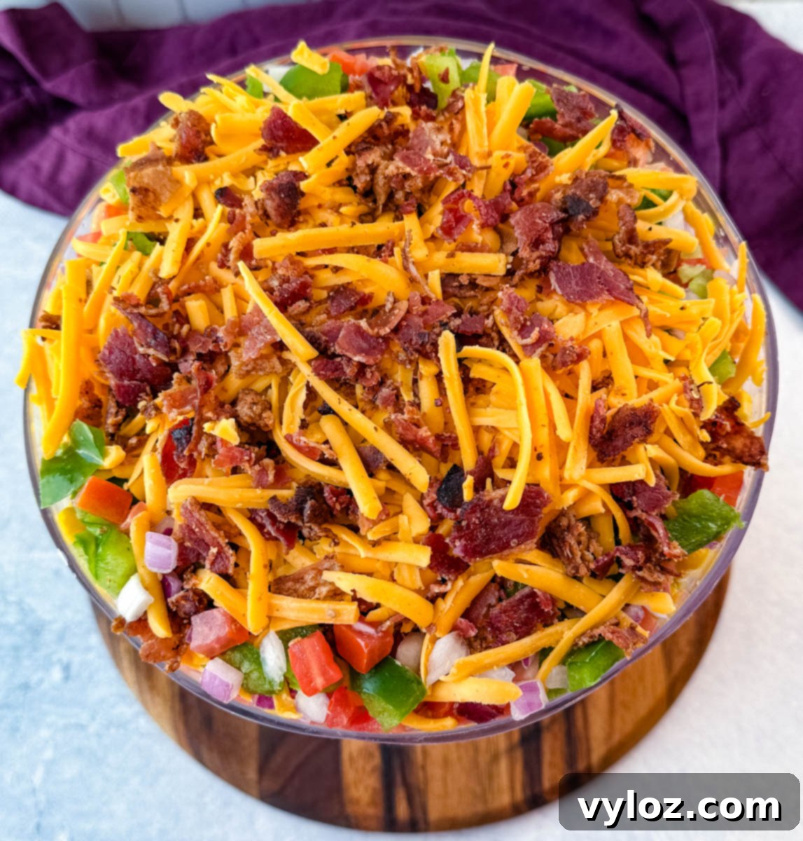 An appetizing overhead close-up of the Southern Cornbread Salad's top layer, richly adorned with shredded cheddar cheese and crumbled bacon, with glimpses of colorful diced green bell peppers, tomatoes, and red onions peeking through.