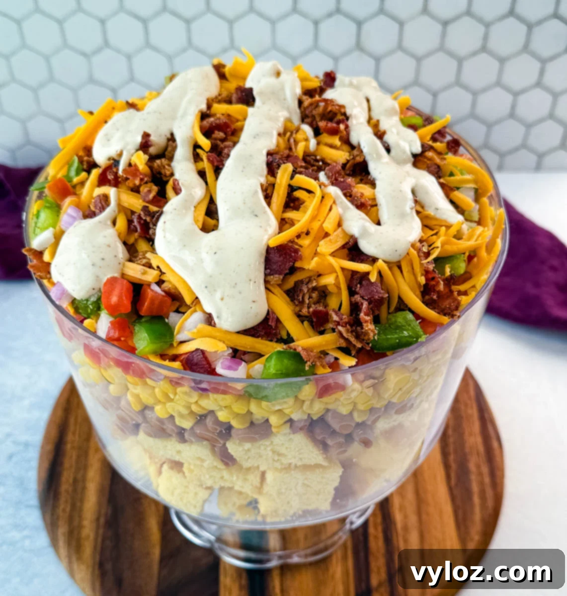 A close-up view of the top layer of the Southern Cornbread Salad, showcasing a rich blend of shredded cheddar, crispy crumbled bacon, and a generous drizzle of thick, creamy ranch dressing, with flecks of green bell peppers, tomatoes, and red onions visible beneath.