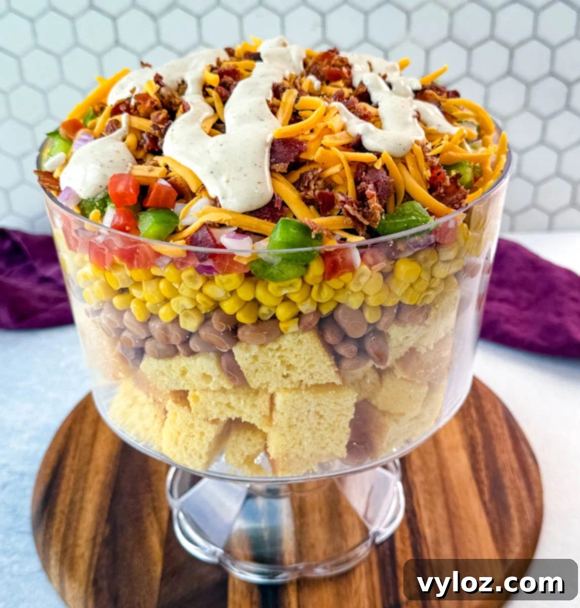 A stunning trifle bowl, elegantly layered with golden cubed cornbread, hearty pinto beans, sweet corn, a colorful array of diced vegetables, sharp shredded cheddar, crispy crumbled bacon, and a final tempting drizzle of creamy ranch dressing on top.