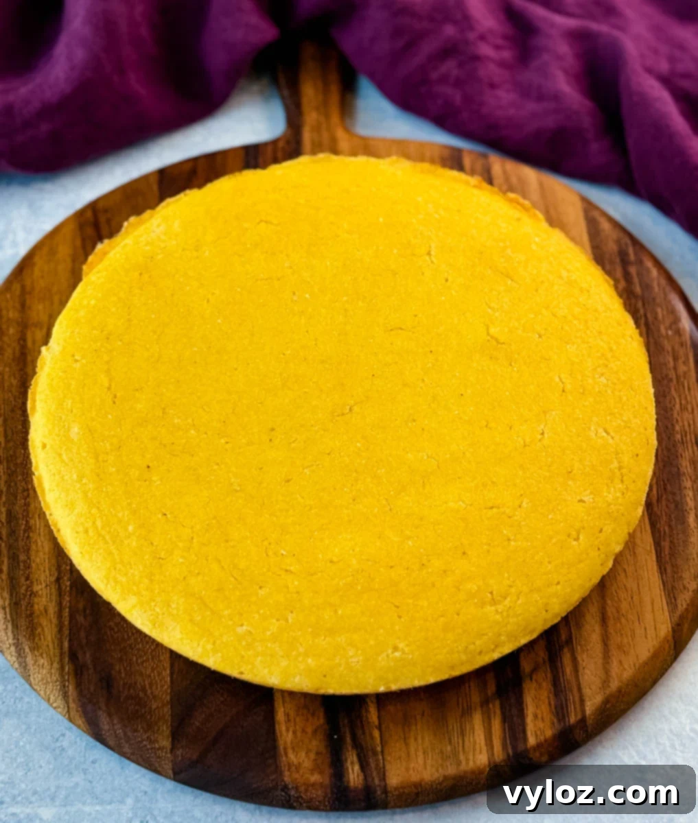 A perfectly golden, round loaf of freshly baked cornbread sits on a rustic wooden board, its inviting aroma promising warmth and comfort, poised for slicing and preparation.