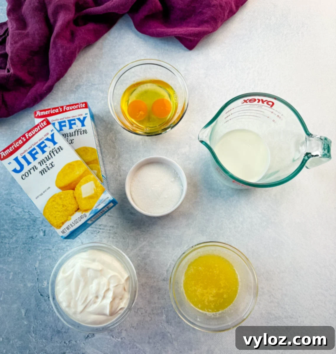 Overhead view of Jiffy cornbread ingredients on a light gray surface, including two boxes of Jiffy corn muffin mix, two cracked eggs in a glass bowl, a measuring cup of milk, a small bowl of sugar, a bowl of sour cream, and a bowl of melted butter, with a purple cloth in the background.