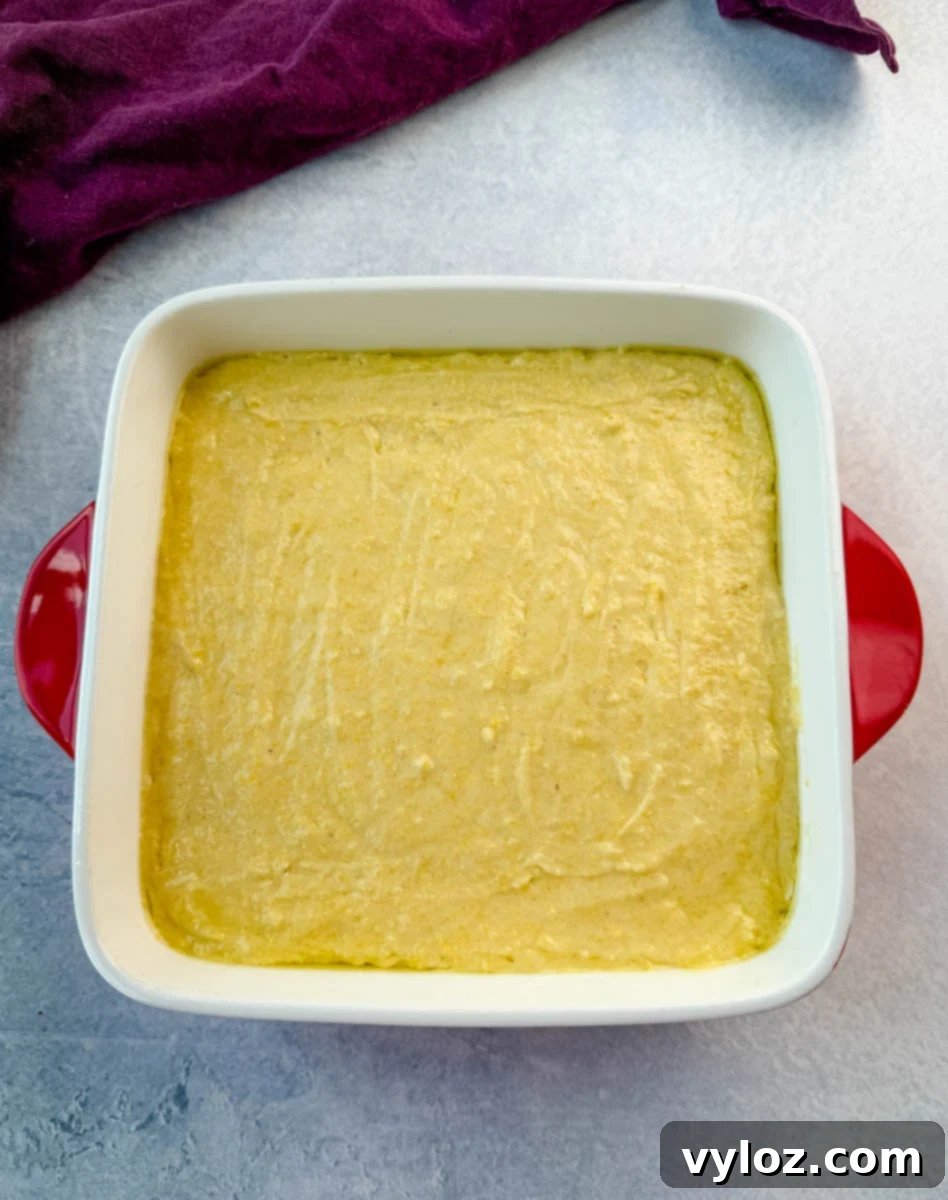 Unbaked cornbread batter spread evenly in a square white baking dish with red handles, ready for the oven, purple cloth in the background.