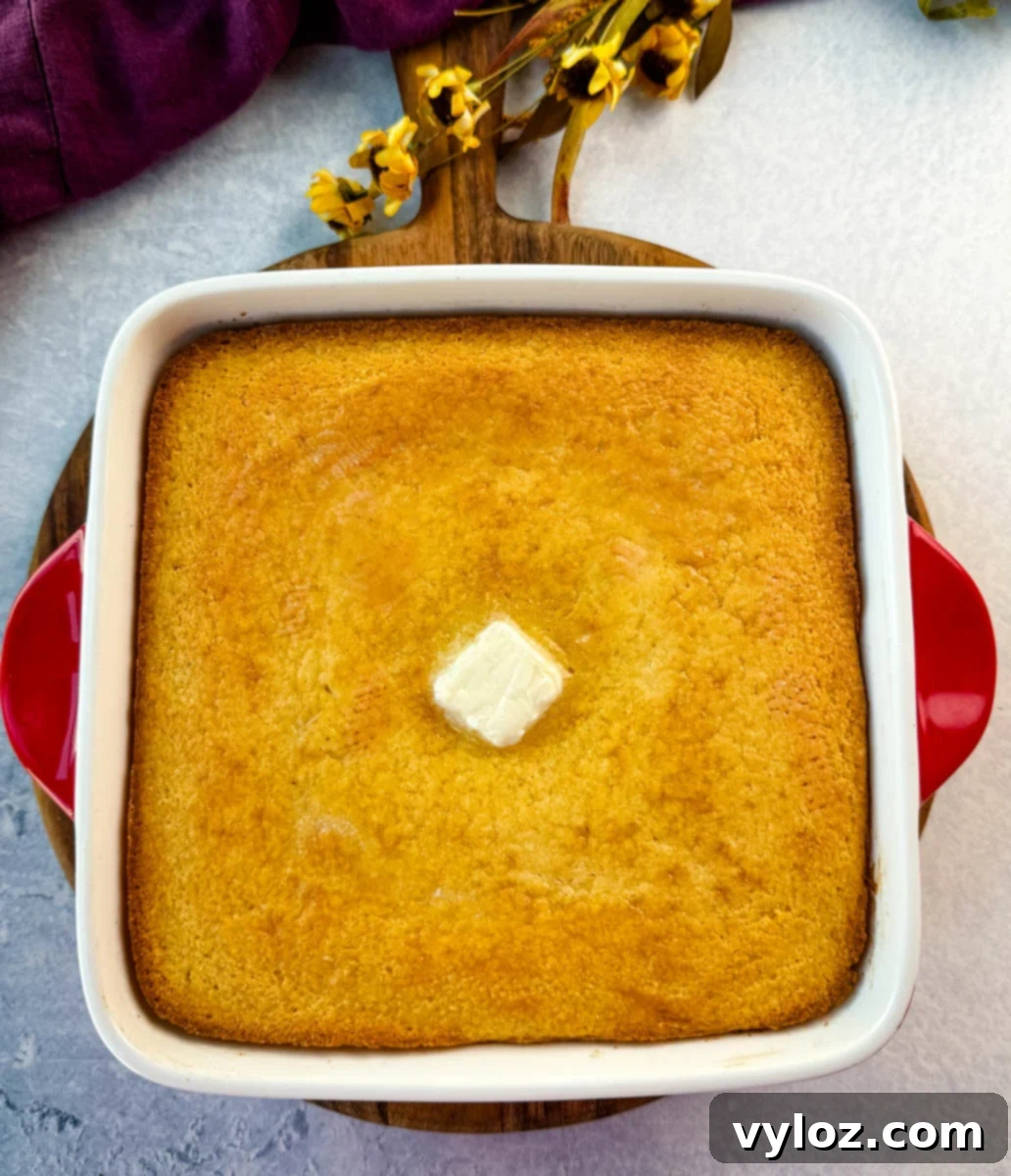 Freshly baked golden cornbread in a square white baking dish with red handles, topped with a pat of butter in the center.