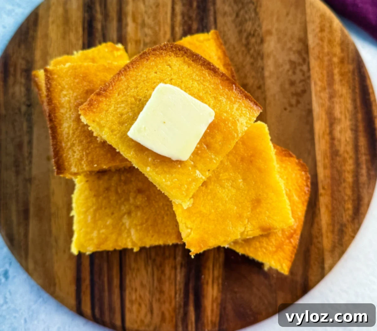Overhead view of stacked cornbread squares on a round wooden board, the top piece topped with a square of butter.