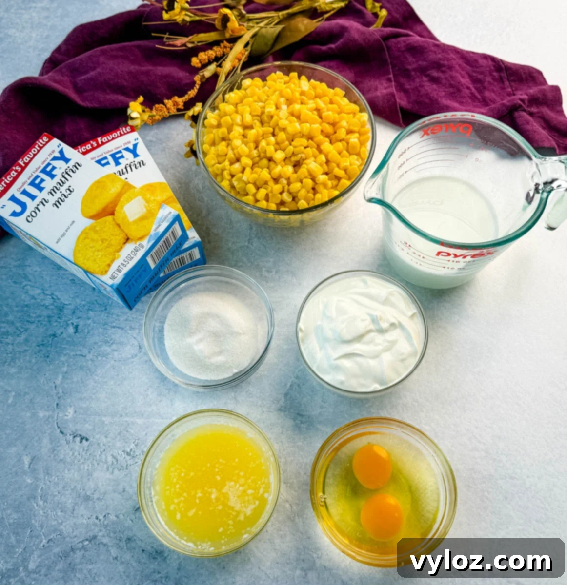 Ingredients for Jiffy corn casserole arranged on a light gray surface, including Jiffy corn muffin mix boxes, a bowl of corn kernels, sugar, sour cream, melted butter, eggs, and milk, with a purple cloth in the background.