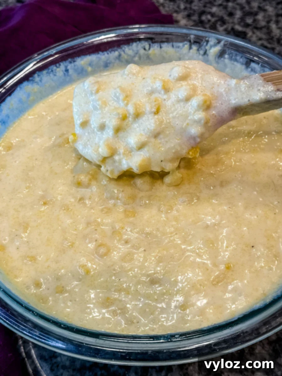 Glass mixing bowl filled with Jiffy corn casserole batter, corn kernels visible, with a wooden spoon lifting a portion.