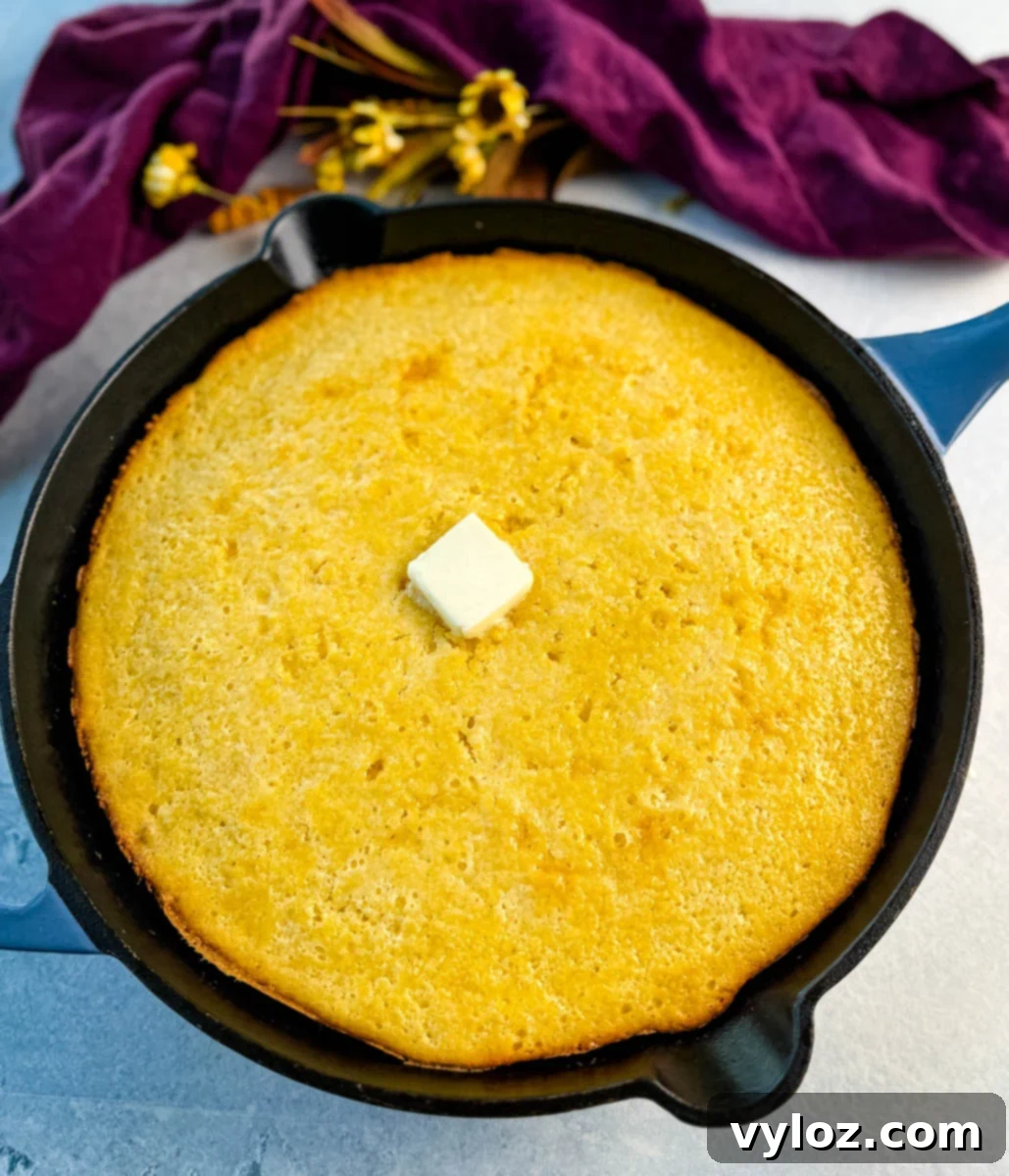 Whole baked Jiffy corn casserole in a black cast iron skillet, topped with a pat of butter in the center, purple cloth and flowers in the background