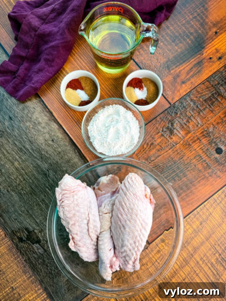Raw turkey wings in a glass bowl surrounded by small bowls of flour and seasoning blends, plus a measuring cup of oil on a wooden surface with a purple cloth.