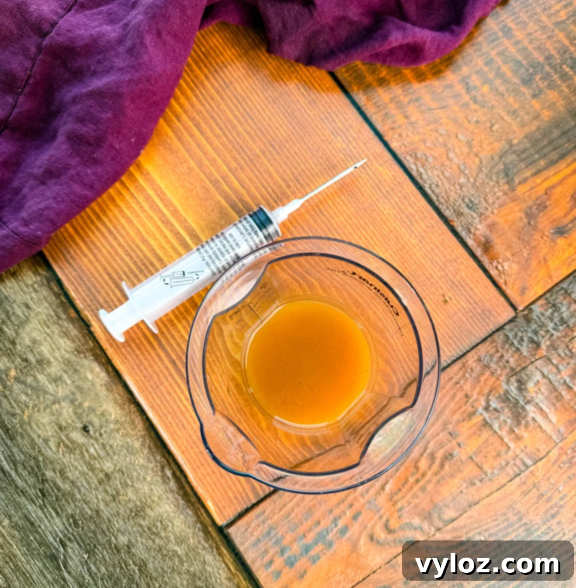 Measuring cup with seasoned injectable marinade and a marinade syringe on a wooden surface with a purple cloth.
