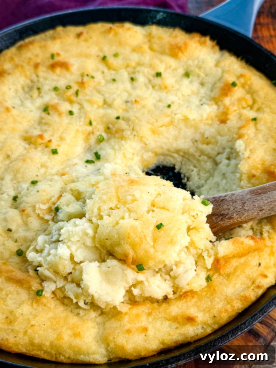 Close-up of a cast-iron skillet filled with golden, fluffy baked mashed potatoes topped with chives, with a wooden spoon scooping out a serving.