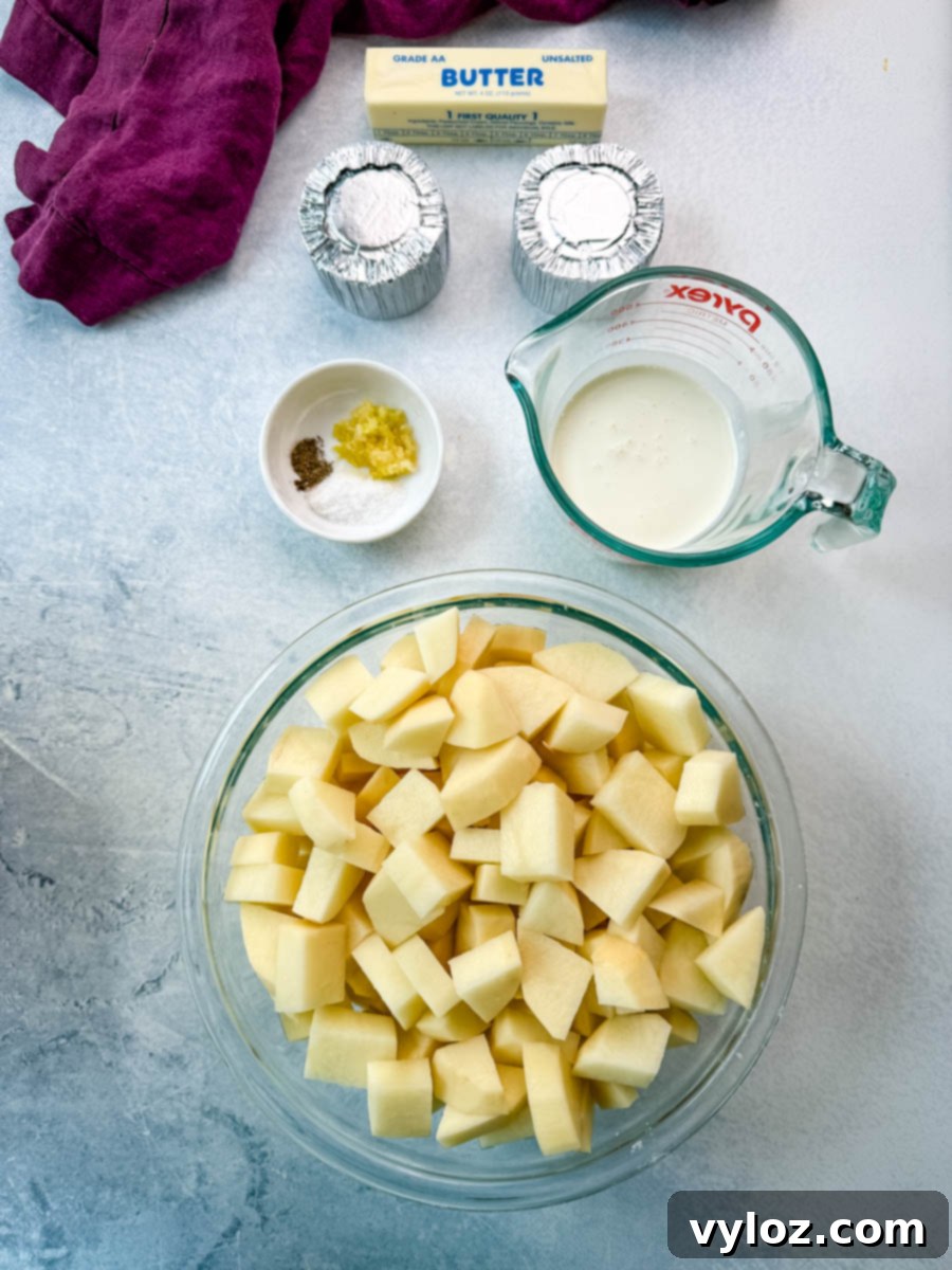 Overhead shot of peeled and cubed potatoes in a large glass bowl, surrounded by a measuring cup of milk, two foil-wrapped rounds of cheese, a stick of unsalted butter, and a small bowl with minced garlic, salt, and pepper, all arranged on a light countertop with a purple kitchen towel.