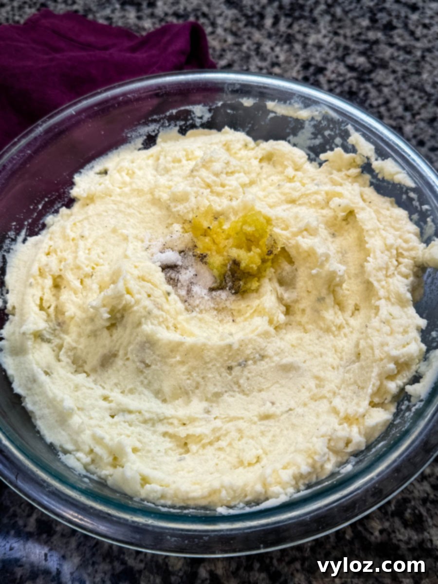 Bowl of smooth mashed potatoes topped with minced garlic, salt, and pepper before mixing.