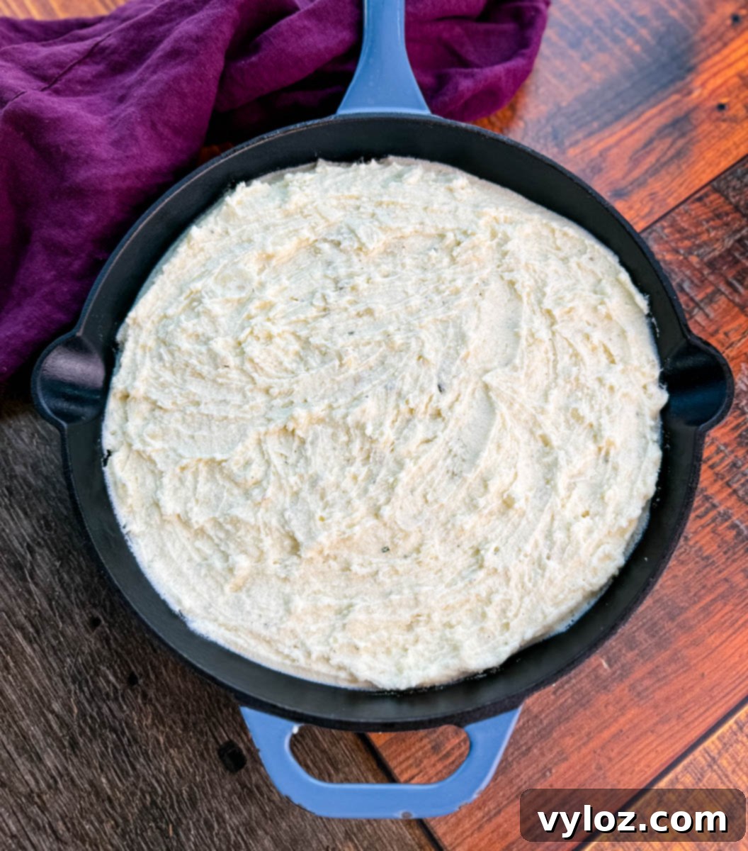 Overhead view of unbaked mashed potatoes spread evenly in a cast-iron skillet, ready for the oven, with a purple cloth in the background.