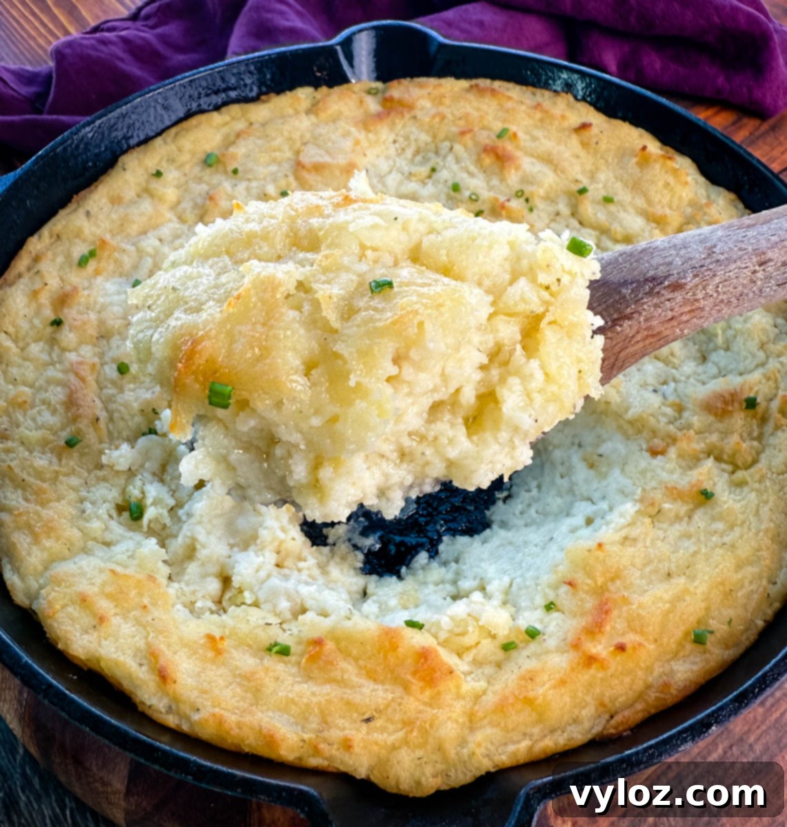 Overhead view of a wooden spoon lifting a large scoop of fluffy, golden baked mashed potatoes from the center of a skillet, garnished with chives.