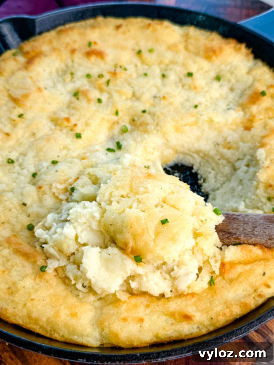 Close-up of a cast-iron skillet filled with golden, fluffy baked mashed potatoes topped with chives, with a wooden spoon scooping out a serving.