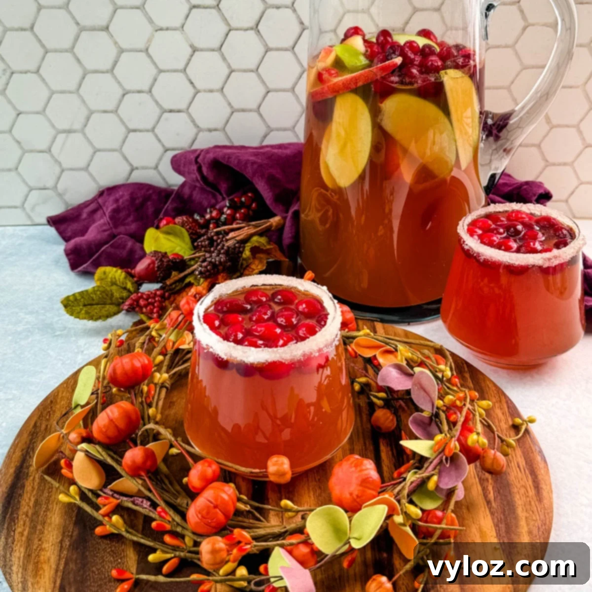 Golden Harvest Punch 2 Pitcher of Thanksgiving punch with apple slices and cranberries, served with two sugared-rim glasses of punch on a round wooden board.