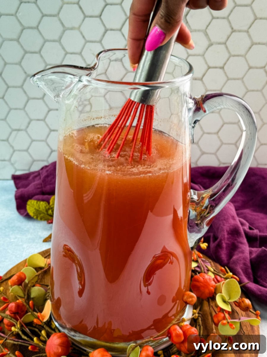 Golden Harvest Punch 6 Hand using a red whisk to stir a pitcher of Thanksgiving punch, blending the spiced mixture