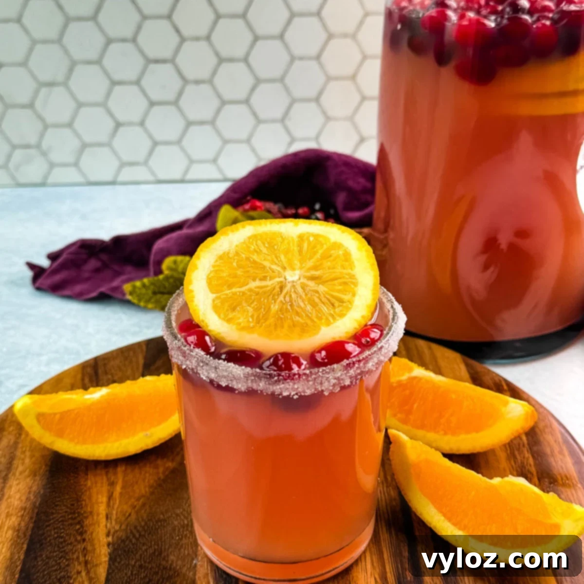 Yuletide Cheer Punch 2 A sparkling glass of non-alcoholic Christmas punch, elegantly garnished with sugared cranberries and a vibrant orange slice. A large pitcher of the festive punch sits invitingly in the soft-focus background, ready for guests.