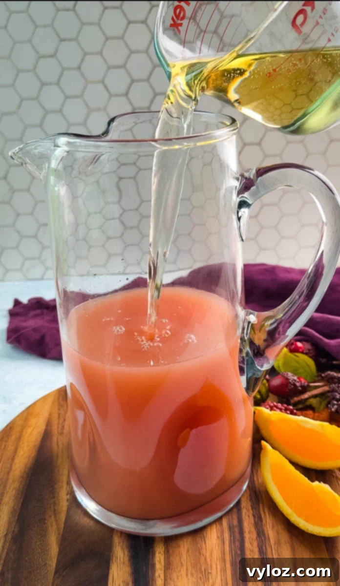 Yuletide Cheer Punch 7 Effervescent lemon-lime soda or ginger ale being carefully poured into a pitcher filled with the pre-mixed punch base, creating a refreshing bubbly top layer. The pitcher rests on a rustic wooden board, accompanied by fresh orange wedges for garnish.