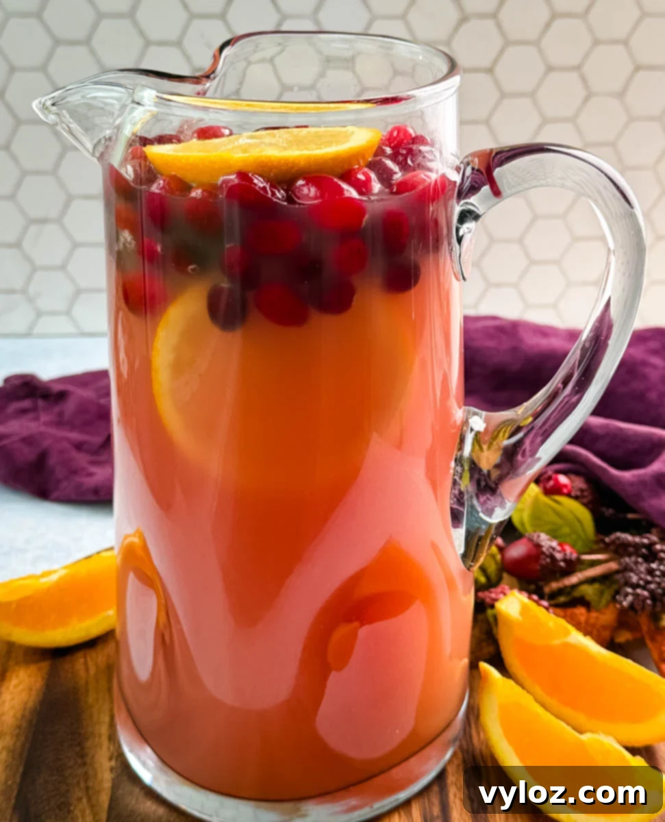 Yuletide Cheer Punch 8 A stunning glass pitcher filled with the completed red holiday punch, beautifully garnished with floating fresh cranberries and delicate orange slices. It is artfully arranged on a wooden board alongside additional fresh orange wedges, ready for serving.