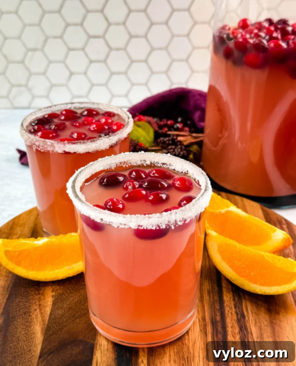 Yuletide Cheer Punch 10 Two elegantly prepared glasses of non-alcoholic Christmas punch, each featuring a sugared rim and garnished with fresh cranberries, positioned in front of a substantial pitcher of the festive beverage.