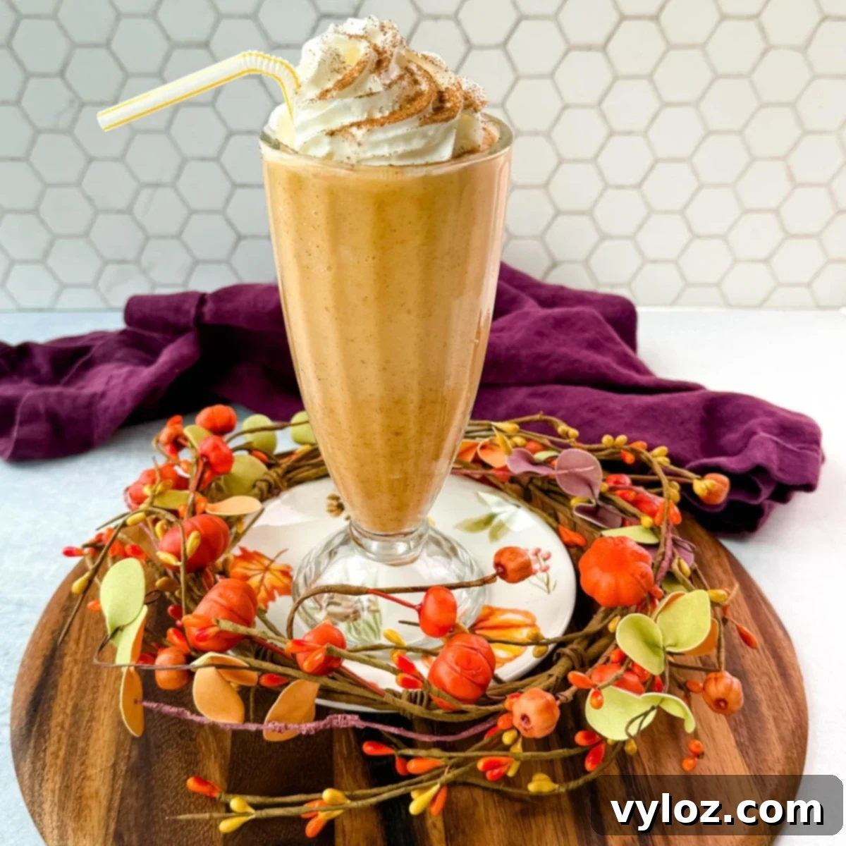 Ninja Slushi's Autumn Spice Elixir 2 A tall glass filled with pumpkin spice milkshake topped with whipped cream and sprinkled with cinnamon, sitting on a fall-themed wooden tray decorated with small pumpkins and leaves.
