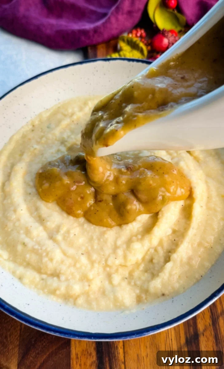 Golden turkey giblet gravy being poured generously from a white gravy boat over a bowl of creamy grits.