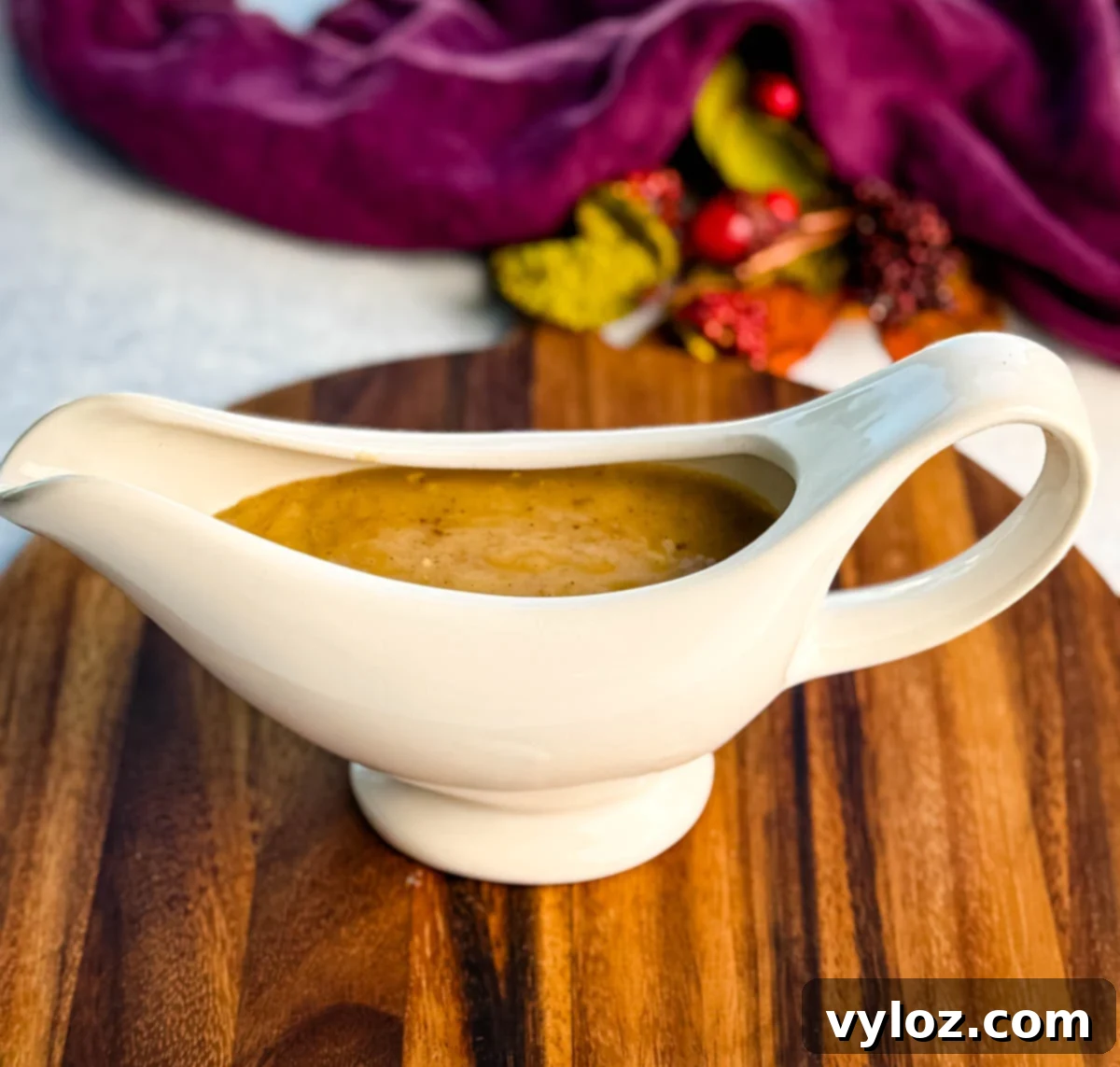White ceramic gravy boat filled with rich, homemade giblet gravy sitting on a wooden board with a purple napkin and fall décor in the background.