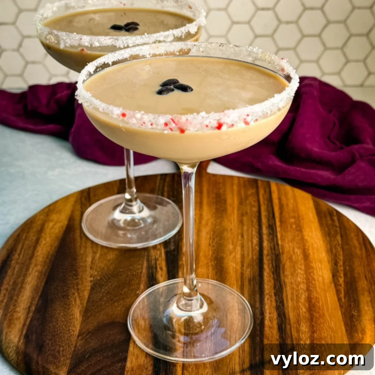 Side view of two peppermint espresso martinis in coupe glasses with crushed peppermint rims and coffee bean garnish, set on a round wooden board with a hexagon tile backsplash.