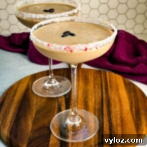 Side view of two peppermint espresso martinis in coupe glasses with crushed peppermint rims and coffee bean garnish, set on a round wooden board with a hexagon tile backsplash.