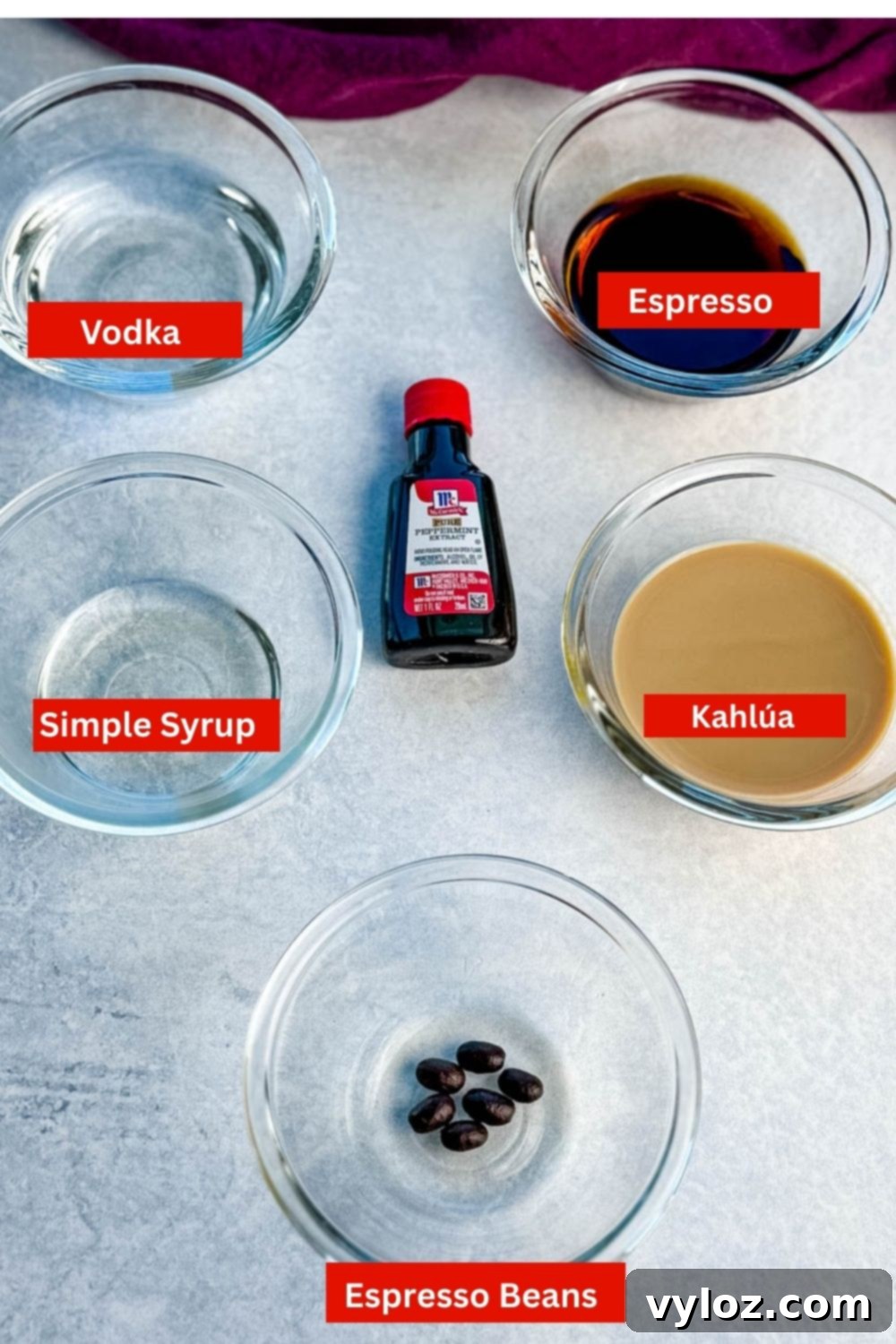 Small glass bowls with ingredients for peppermint espresso martinis including vodka, coffee liqueur, Baileys, peppermint extract, and coffee beans arranged neatly on a gray surface.