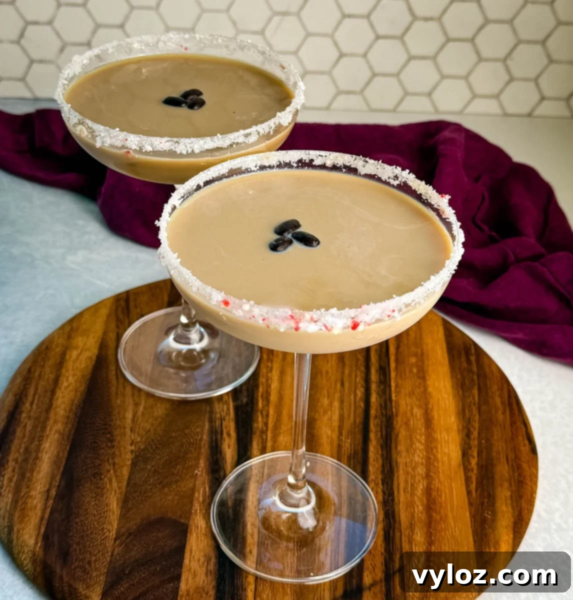 Two coupe glasses filled with creamy peppermint espresso martinis, each rimmed with crushed peppermint and garnished with three coffee beans, displayed on a wooden board with a deep purple cloth in the background.