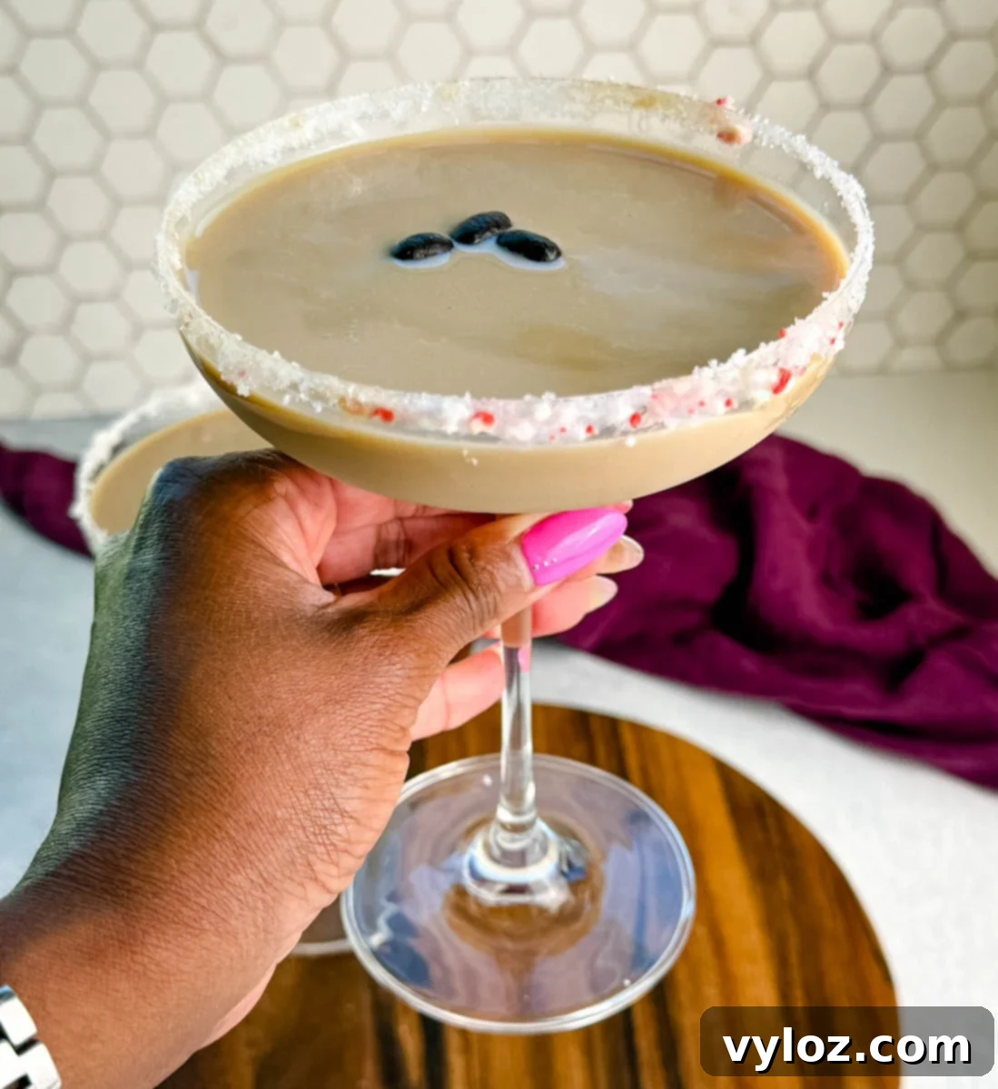 Hand holding a coupe glass with a finished peppermint espresso martini, rimmed with crushed peppermint and garnished with coffee beans, with another glass blurred in the background.